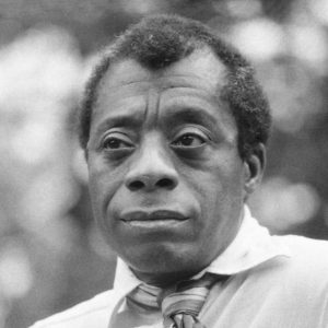A black and white portrait photo of James Baldwin, taken from the shoulders up. The background is out of focus and highlights Baldwin, who appears to be sitting down and wearing a light colored button down shirt and a striped tie.