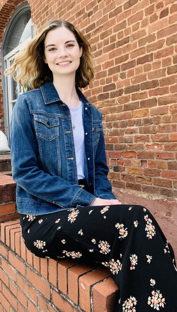 Ariana Adams in a jean jacket, white shirt, and floral skirt sitting on a brick retaining wall.