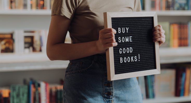 Woman in front of a book shelf holding a sign that says, "Buy some good books!"